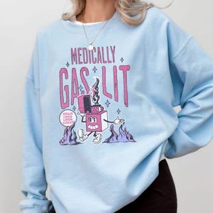 Pode incluir: Camisola azul claro com um gr&aacute;fico de um isqueiro de desenho animado com o texto "Medically Gaslit" e "Your Labs Look Great!" em rosa e preto.