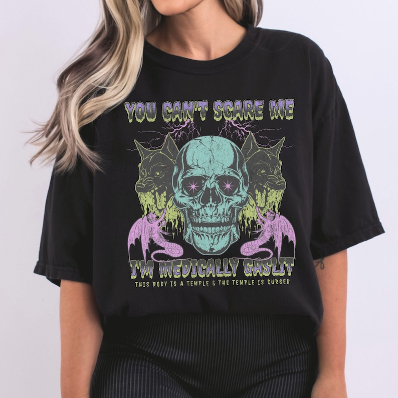 May include: Black t-shirt with a teal skull graphic, flanked by two snarling dog-like creatures and two purple bat-like figures. The text "YOU CAN'T SCARE ME" and "I'M MEDICALLY GASLIT" are printed above and below the skull.