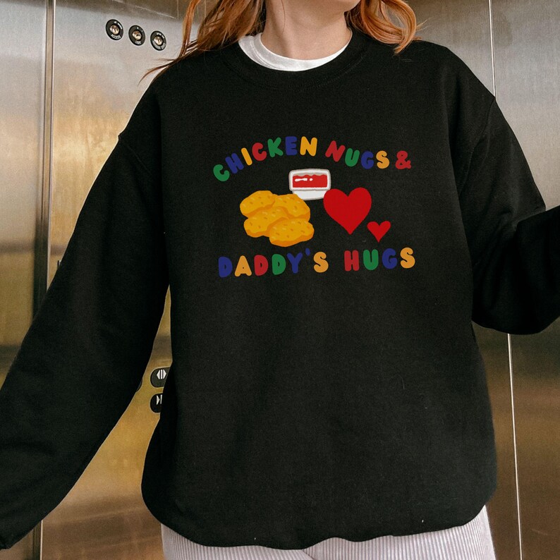 May include: Black sweatshirt with the text "CHICKEN NUGS & DADDY'S HUGS" in rainbow colors. The design includes illustrations of chicken nuggets, a container of dipping sauce, and red hearts. The sweatshirt has a crew neck and long sleeves.
