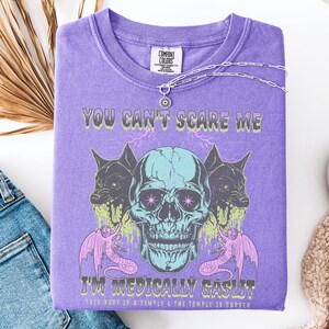 May include: A purple Comfort Colors t-shirt with a graphic design featuring a teal skull, two black dog-like creatures, and pink serpent-like creatures. The text reads "YOU CAN'T SCARE ME" and "I'M MEDICALLY GASLIT".