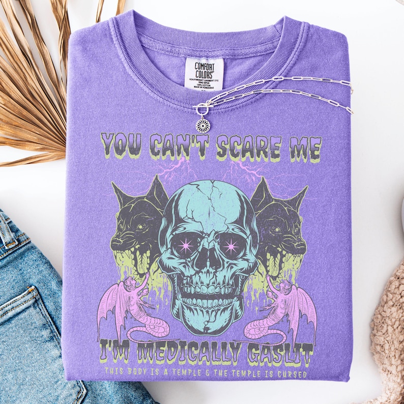 May include: A purple Comfort Colors t-shirt with a graphic design featuring a teal skull, two black dog-like creatures, and pink serpent-like creatures. The text reads "YOU CAN'T SCARE ME" and "I'M MEDICALLY GASLIT".