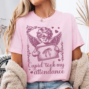 May include: Pink t-shirt featuring a Cupid graphic with heart-shaped sunglasses and wings, holding a sign with a bow. The text "Cupid took my attendance" is below the graphic. The shirt is a light pink color.