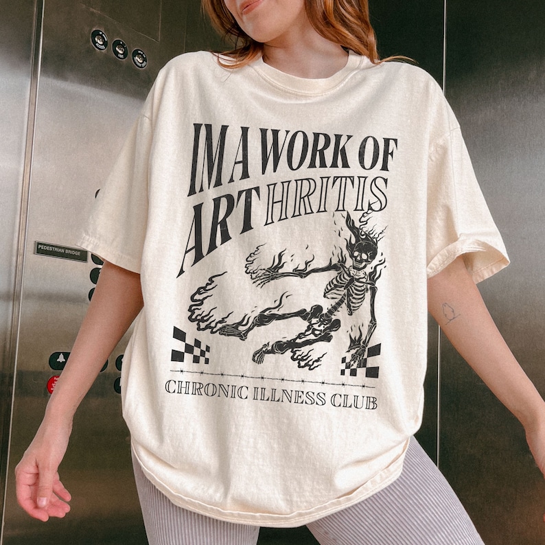 Pu&ograve; includere: Una maglietta color crema con una grafica nera di uno scheletro in fiamme. Il testo sulla maglietta recita "I'm a work of arthritis" e "Chronic Illness Club".