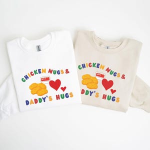 May include: Two long-sleeved sweatshirts, one white and one beige, with a colorful graphic. The graphic reads "CHICKEN NUGS & DADDY'S HUGS" with images of chicken nuggets, a heart, and a container of dipping sauce.