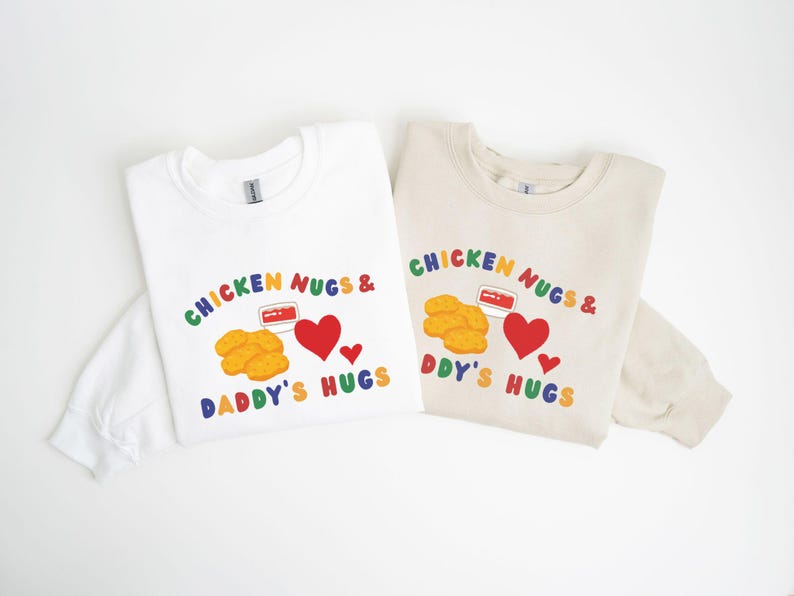 May include: Two long-sleeved sweatshirts, one white and one beige, with a colorful graphic. The graphic reads "CHICKEN NUGS & DADDY'S HUGS" with images of chicken nuggets, a heart, and a container of dipping sauce.