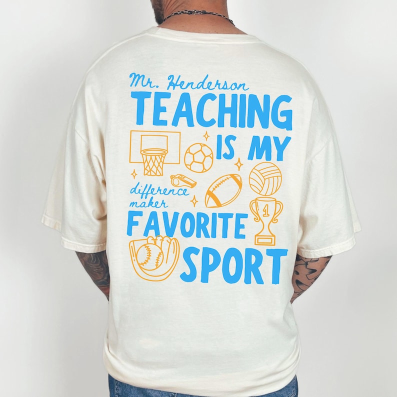 May include: Cream-colored t-shirt with blue and orange text and graphics. The shirt says "Mr. Henderson TEACHING IS MY FAVORITE SPORT" with sports-related illustrations. A basketball hoop, soccer ball, football, volleyball, baseball, and trophy are shown.