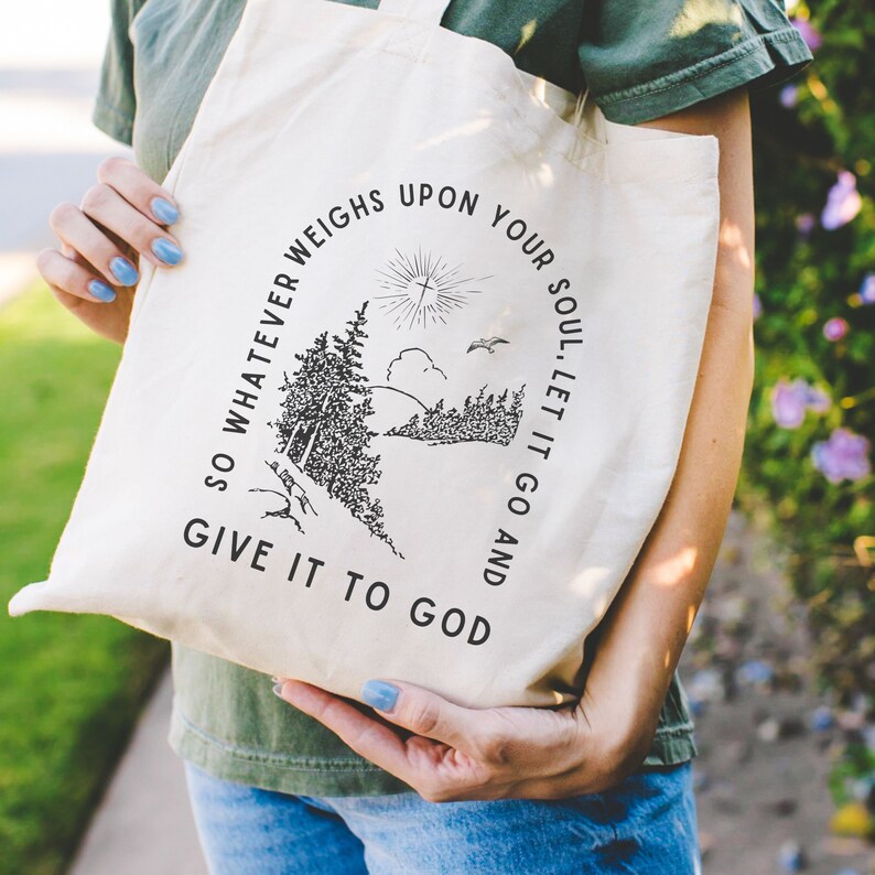 May include: A white canvas tote bag with black text and a graphic of a forest scene. The text reads "So whatever weighs upon your soul, let it go and give it to God."