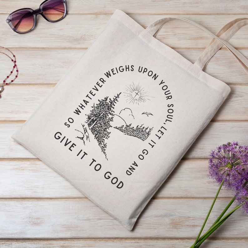 May include: A white canvas tote bag with black text that reads "SO WHATEVER WEIGHS UPON YOUR SOUL, LET IT GO AND GIVE IT TO GOD". The text is in a circle around a black and white illustration of a forest scene with a bird flying overhead.