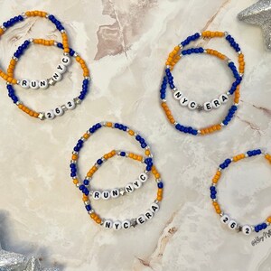 May include: Four beaded bracelets with alternating blue and orange beads. The bracelets feature white letter beads spelling out "RUN NYC", "NYC ERA", and "262".