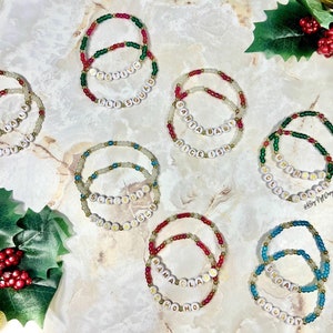 May include: A collection of beaded bracelets with holiday-themed words. The bracelets feature red, green, gold, and clear beads. The words include "Jolly", "Festive", "Santa Baby", "Nice-ish", "Ho Ho Ho", and "Snowy".