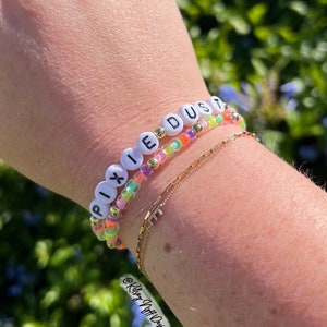 May include: A close-up of a person's wrist wearing two beaded bracelets. The top bracelet spells out "PIXIE DUST" in white letters on colorful beads. The bottom bracelet is a simple gold chain. The bracelets are against a background of green foliage.