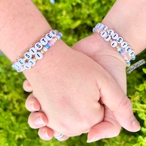 May include: Two wrists with beaded bracelets that spell out "Little Sister" in white letters on a pink, blue, and white background. The bracelets are on a gold chain.