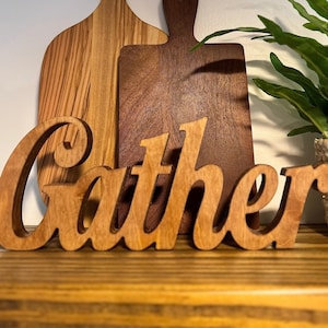 May include: Wooden 'Gather' sign in a cursive font, placed in front of two wooden cutting boards. The cutting boards are different shades of brown. A potted plant in a woven basket is on the right side of the image.