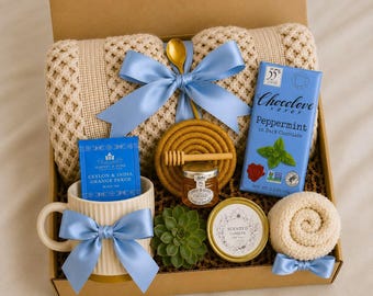 May include: A gift box containing a beige knit blanket with a blue ribbon, a mug with a blue ribbon, a blue box of Harney & Sons tea, a bar of Chocolove peppermint dark chocolate, a jar of honey with a wooden dipper, a succulent, and a candle.