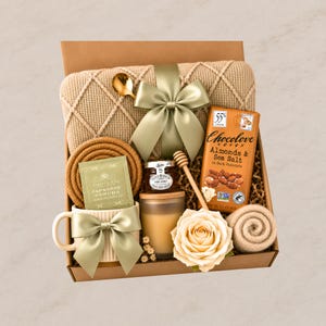 May include: A gift box containing a beige knit blanket with a sage green ribbon, a mug with a sage green bow, a bar of Chocolove Almonds & Sea Salt dark chocolate, a jar of honey with a wooden dipper, a tea sachet, a candle, and a rose.