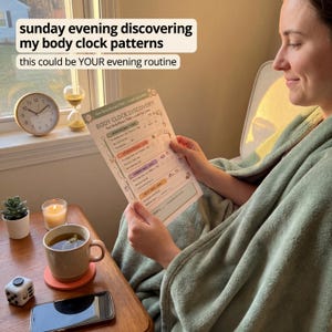 Woman curled up in reading chair wrapped in sage green throw blanket reading Body Clock Discovery page with sunrise to moon timeline blocks with candle tea mug hourglass and clock nearby and Instagram style caption reading sunday evening discovering