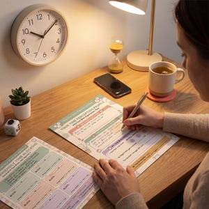 Person at cozy desk actively writing on Your Time Perception Profile worksheet page with pen in hand and another Time Blocking page nearby surrounded by wall clock hourglass warm desk lamp tea on coral coaster succulent and fidget cube in warm ambien
