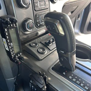 May include: A car's interior console with a black gear stick and a radio communication device. The console includes buttons for media, climate control, and other functions. The gear stick is in the 'P' position. The overall colour scheme is grey and black.