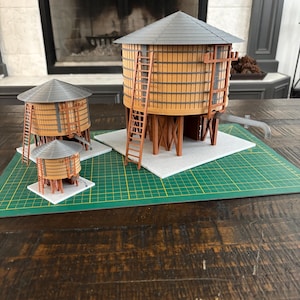 Könnte beinhalten: Eine Sammlung von Miniatur-Wasserturmmodellen. Die Türme sind aus Holz gefertigt und haben eine zylindrische Form mit einem grauen Dach. Jeder Turm hat eine Leiter und ist auf einer quadratischen Basis montiert. Die Modelle werden auf einer grünen Schneidematte präsentiert.
