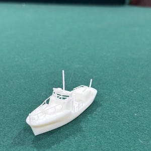 Puede incluir: Un pequeño modelo de barco blanco con detalles como cabina, barandillas y mástiles. El barco se encuentra sobre un fondo verde, lo que sugiere un tema náutico o un interés por el modelismo. El modelo es una réplica en miniatura de un barco.