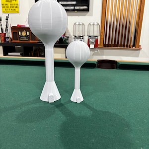 Puede incluir: Dos modelos blancos impresos en 3D de torres de agua de diferentes tamaños, de pie sobre una superficie de fieltro verde. Las torres tienen una parte superior bulbosa, un tallo cilíndrico y una base acampanada. La torre más grande mide unos 30 cm de altura.