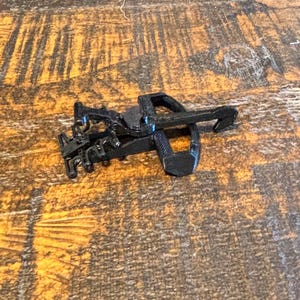 May include: A small, black, toy crossbow with a detailed design. The crossbow is made of plastic and has a trigger and a bow. The toy is lying on a wooden surface.