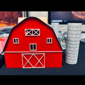 May include: A miniature red barn with white trim and a black roof. The barn features a white door and window details. A gray silo is positioned next to the barn, creating a farm scene.