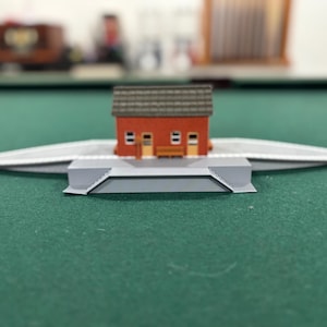 May include: A miniature model of a train station. The station has a red brick exterior, a gray roof, and a gray platform. The model is set on a green surface.