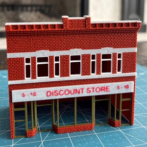 May include: A miniature red brick building with white trim and windows. The sign reads "DISCOUNT STORE 5-10" in red letters. The building has a detailed facade and is likely a model or a toy.