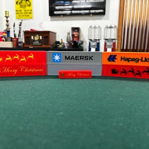 May include: Miniature shipping container models in red, grey, and orange, with the logos of Maersk and Hapag-Lloyd. The red containers have "Merry Christmas" text and a reindeer sleigh design. The containers are on a green surface.