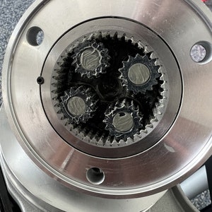 May include: Close-up of a silver metal gear system with three interlocking gears. The gears are visible through a circular opening in a larger metal plate.