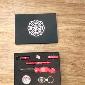 May include: A black gift box with a white fire department logo on the lid. The box is open and contains a red pocket knife, a red flashlight, a small American flag, and a keychain with a fire department logo.