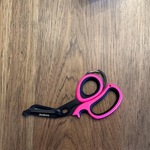 May include: Black and pink medical shears with a keyring attachment. The shears have a black blade and handle with pink finger holes. The name "Andrea" is printed on the blade.