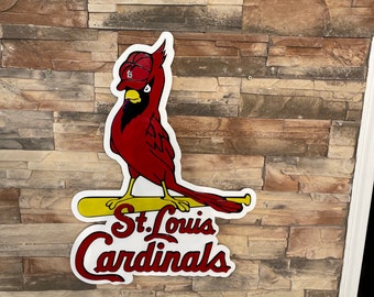 May include: A red and white St. Louis Cardinals sign featuring a cartoon cardinal bird wearing a baseball cap. The bird is perched on a yellow baseball bat, with the team name in red script below.