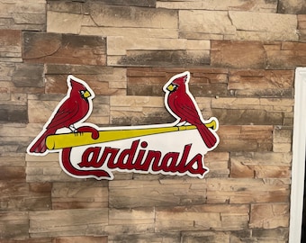 May include: A white sign with the word "Cardinals" in red lettering. Two red cardinals are perched on a yellow baseball bat.