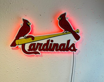 May include: A neon sign featuring two red cardinal birds perched on a yellow baseball bat. The word "Cardinals" is written in red script below the birds. The sign has a red neon glow.
