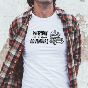 May include: White t-shirt with a black graphic of a camper van and the text "EVERYDAY is a new ADVENTURE". The person is wearing a red and black plaid shirt.