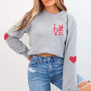 May include: A gray sweatshirt with a red skeleton hand design that spells out "LOVE". The sweatshirt has red heart patches on the sleeves.