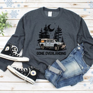 May include: A dark gray sweatshirt with a graphic of a truck with a rooftop tent driving through a forest at night. The text "Gone Overlanding" is printed below the truck. The sweatshirt is laid out on a wooden surface with a pair of black high-top sneakers and blue jeans.