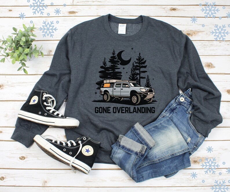 May include: A dark gray sweatshirt with a graphic of a truck with a rooftop tent driving through a forest at night. The text "Gone Overlanding" is printed below the truck. The sweatshirt is laid out on a wooden surface with a pair of black high-top sneakers and blue jeans.