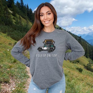 May include: A gray long-sleeved shirt with a cartoon illustration of a car with a tent on top. The text "I SLEEP ON TOP" is printed below the illustration.