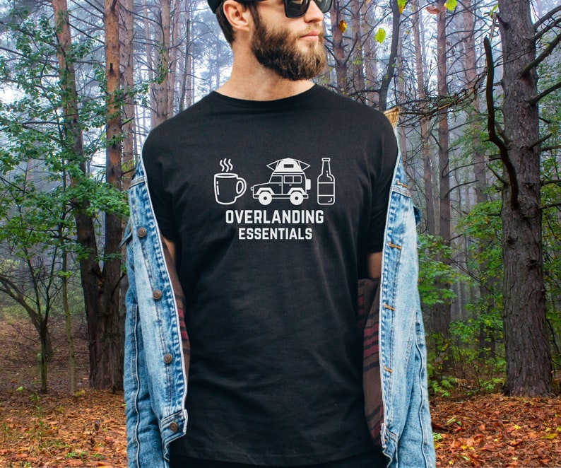 Puede incluir: Camiseta negra con un gr&aacute;fico blanco de un coche, una tienda de campa&ntilde;a, una taza de caf&eacute; y una botella de cerveza. El texto "Overlanding Essentials" est&aacute; debajo del gr&aacute;fico.