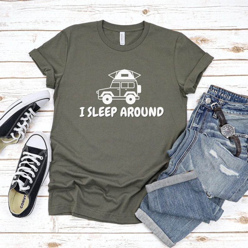 Pu&ograve; includere: Una maglietta verde con una grafica bianca di un'auto con una tenda sul tetto. Il testo "I SLEEP AROUND" &egrave; stampato sotto l'auto.