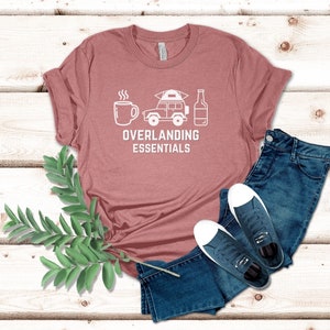Puede incluir: Una camiseta rosa con un gr&aacute;fico blanco de una taza de caf&eacute;, un jeep con una tienda de campa&ntilde;a en la parte superior y una botella de cerveza. El texto "Overlanding Essentials" est&aacute; debajo del gr&aacute;fico.