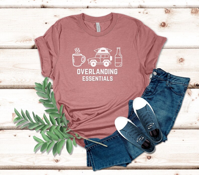 Puede incluir: Una camiseta rosa con un gr&aacute;fico blanco de una taza de caf&eacute;, un jeep con una tienda de campa&ntilde;a en la parte superior y una botella de cerveza. El texto "Overlanding Essentials" est&aacute; debajo del gr&aacute;fico.