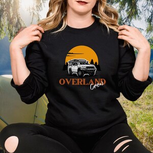 May include: Black sweatshirt featuring an orange sunset graphic and a white truck with a roof rack. The text "Overland Crew" is printed in orange below the truck.