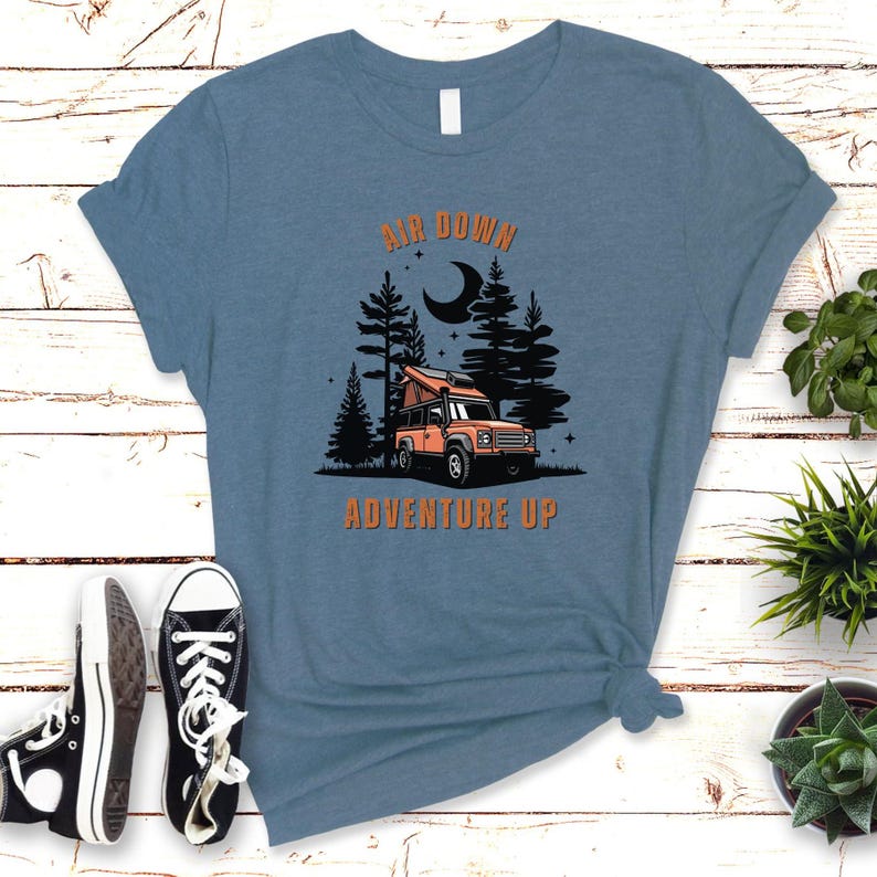 May include: A blue t-shirt with the text "Air Down Adventure Up" in orange. The design features a cartoon orange jeep in a forest setting with a crescent moon in the sky.