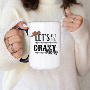 May include: White ceramic mug with a black interior and handle, featuring the text "LET'S PUT THE CRAZY AWAY" in black, with a brown cowboy hat graphic. The mug is held by a person wearing a white sweater.