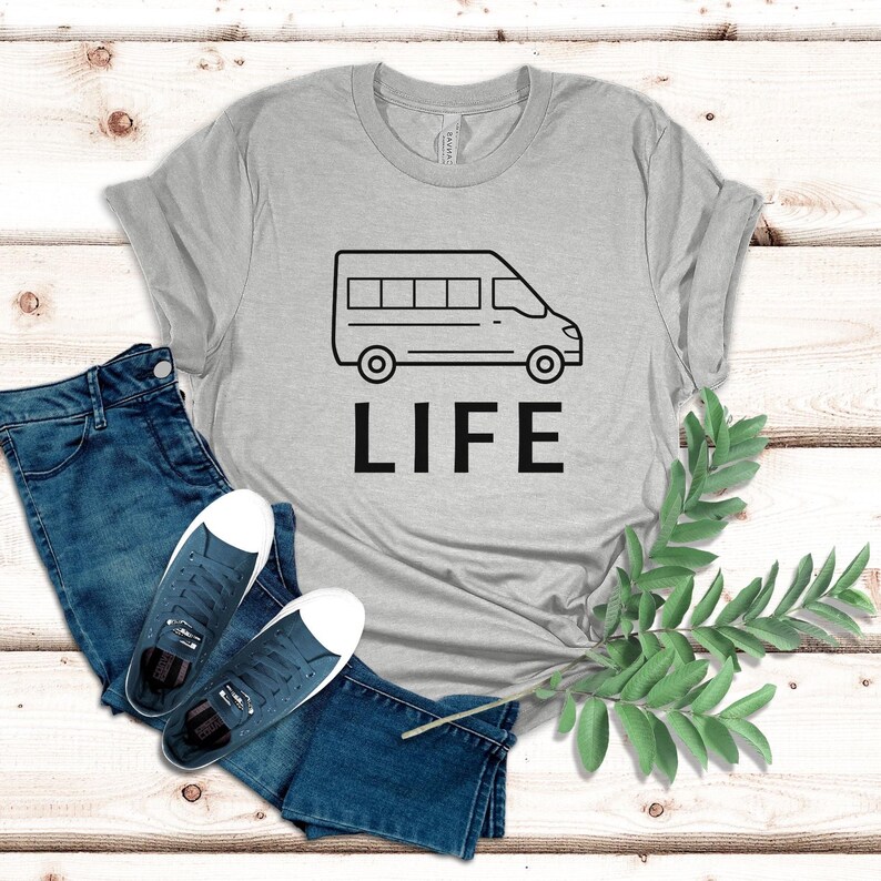 Op de afbeelding: Een grijs T-shirt met een zwarte omtrek van een bestelwagen en het woord "LIFE" erop gedrukt. Het T-shirt ligt op een houten oppervlak met een blauwe spijkerbroek en een paar witte sneakers.