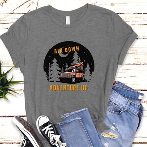 Puede incluir: Camiseta gris con un gr&aacute;fico de un coche en un bosque de noche. El texto "Air Down Adventure Up" est&aacute; impreso en la camiseta.